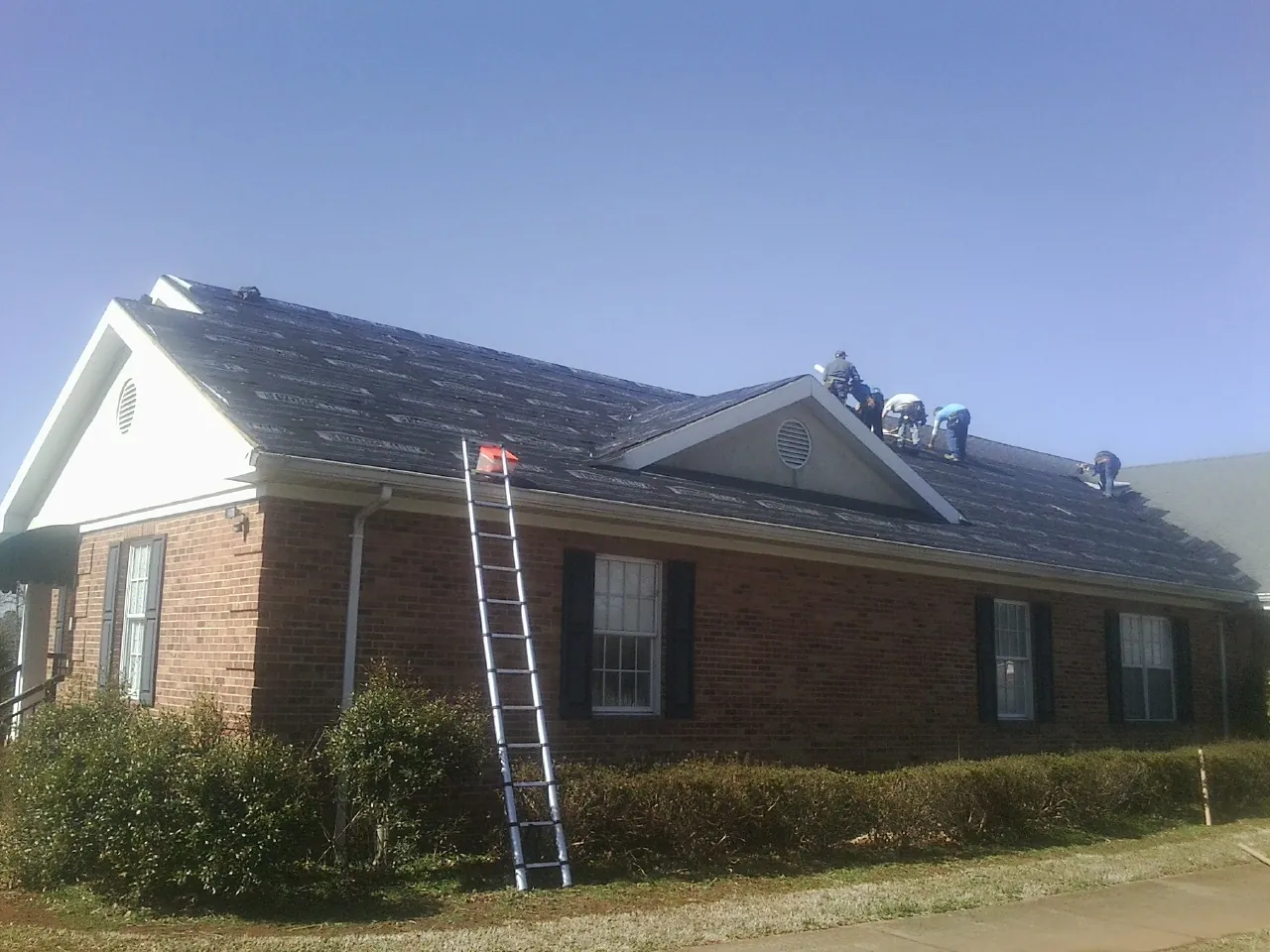 Crew tearing shingles off a brick ranch roof with a ladder set — Emergency Roof Repair work by our crew in East Baldwin