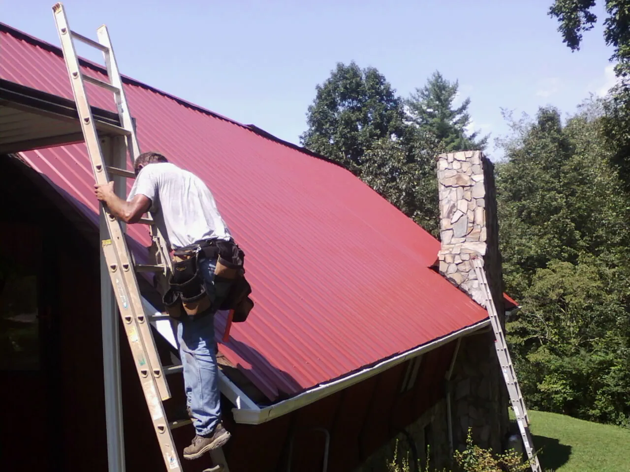 Roofer on a ladder beside a red metal roof and stone chimney — Soffit & Fascia Repair by Roofing Brothers in East Baldwin